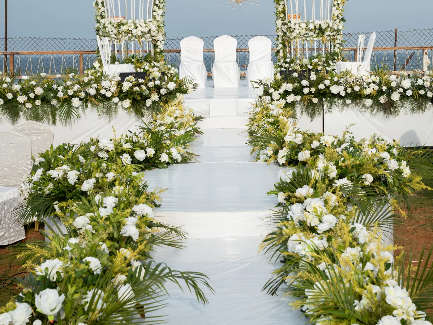 Walkway edged with white roses, lilies and palm leaves leading to the wedding stage