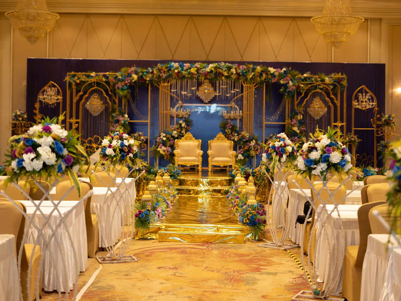 Reception hall with a blue and gold stage, floral stands along a gold aisle and tables set either side